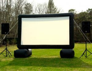 An inflatable outdoor movie screen is set up on a grassy lawn, flanked by two large speakers on stands, with trees and bushes in the background—perfect for party rentals Long Island events.