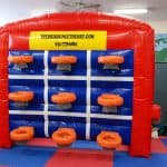 An inflatable climbing wall with blue and white grid lines, orange handholds, and a yellow sign reading "THEBIGBOUNCETHEORY.COM 631-778-6861" on top, perfect for party rentals Long Island, stands on a colorful padded floor.