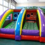 A colorful inflatable football toss game from party rentals Long Island features two lanes with green field designs and circular target holes, set up indoors next to large windows and a mural wall.