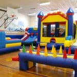 A school gym set up for indoor play features colorful inflatable obstacle courses and a bounce house Long Island kids love, including a slide, climbing wall, and traffic cones. Basketball hoops and a mural add extra fun to this party rentals Long Island setup.