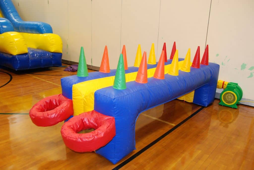 A colorful inflatable game from party rentals Long Island, with red, yellow, and blue sections, two red baskets at one end, and rows of orange, green, and yellow cones on top, set on a gym floor with another bounce house Long Island in the background.