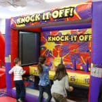 Three children toss balls at targets in an inflatable carnival game called "Knock It Off!" from a party rentals Long Island company, while an adult watches nearby. The game features bright colors and numbered targets, perfect for NY events.