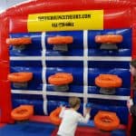 A young boy reaches for basketball hoops on an inflatable game wall while a woman watches nearby at an indoor play center, perfect for party rentals Long Island. The red wall features orange hoops, a blue background, and a yellow company sign at the top.