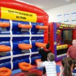 Children play in front of an inflatable game wall with orange targets at a lively NY event. In the background, a “Knock It Off” booth from party rentals Long Island adds to the indoor fun as others gather nearby.
