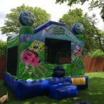 A colorful SpongeBob SquarePants-themed bounce house stands on grass in NY, decorated with images of SpongeBob, Patrick, jellyfish, and sea designs. Trees and a wooden fence are in the background; a small dog is nearby. Perfect for party rentals Long Island!.