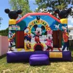 A colorful Mickey Mouse-themed bounce house Long Island sits on grass in a backyard, decorated with images of Mickey, Minnie, Donald, Goofy, and Pluto. Trees, a white fence, and houses complete this perfect NY party rentals Long Island scene.