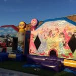 Two colorful inflatable bounce houses are set up outdoors on grass in NY. Perfect for party rentals Long Island, one features Disney princesses and castles, while the other showcases Cars characters with arched entrances and balloons on top.