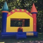 A colorful inflatable bounce house Long Island with red, yellow, blue, and green towers stands on a grassy area under tall trees, with sunlight filtering through the leaves—a perfect spot for NY party rentals Long Island.