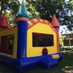 A colorful inflatable bounce house Long Island with red, blue, yellow, and orange castle-like turrets is set up on grass in a shaded backyard area with trees and a house in the background—perfect for party rentals Long Island, NY.