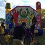 Children and adults stand in line in front of a colorful Yo Gabba Gabba! themed bounce house Long Island at an outdoor event on a sunny day.