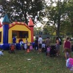Children and adults gather around a colorful bounce house Long Island, NY at a park, with some waiting in line and others playing nearby. Strollers and backpacks are visible on the grass under trees.