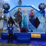 A man stands in front of a large inflatable bounce house Long Island decorated with Disney Frozen characters, including Kristoff, Olaf, Elsa, and Anna, set up outdoors in a NY backyard.