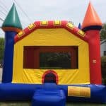 A colorful inflatable bounce house Long Island with red, yellow, blue, and green sections sits on a lawn near a street and houses, featuring towers and an arched entrance—perfect for party rentals in NY.