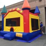 A colorful inflatable bounce house Long Island with red, yellow, and blue castle-like towers is set up outside on a blue tarp near a building. A person stands on the right side adjusting it at this fun NY party scene.