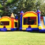 Two colorful inflatable bounce houses sit on a grassy lawn in NY; one features a slide, and both have bright red, yellow, blue, and orange sections—perfect for party rentals Long Island with trees in the background.