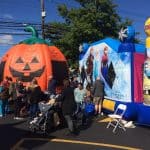 People gather at an outdoor event with large inflatable bounce houses, including a pumpkin and Disney Frozen theme, on a sunny day. Kids and adults enjoy the fun, thanks to party rentals Long Island bringing excitement to the gathering.