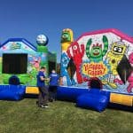 Two colorful inflatable bounce houses on grass—one with SpongeBob SquarePants and the other with Yo Gabba Gabba! characters—showcase fun party rentals Long Island style, as two people in blue shirts stand between them under a clear blue sky.