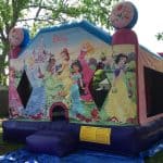 A colorful Disney princess-themed bounce house Long Island sits outdoors on a blue tarp, surrounded by trees and a wooden fence, perfect for party rentals Long Island, NY, and featuring images of several princesses and a castle.