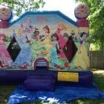 A colorful bounce house Long Island decorated with Disney princesses, including Cinderella, Belle, Ariel, and others, stands on blue tarp in a shaded, grassy backyard with a wooden fence and trees.