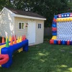 A backyard in NY with a beige shed, an inflatable game table with colorful cones and rings, a large inflatable Plinko-style game, and lush green grass—perfect for party rentals Long Island style, surrounded by trees.