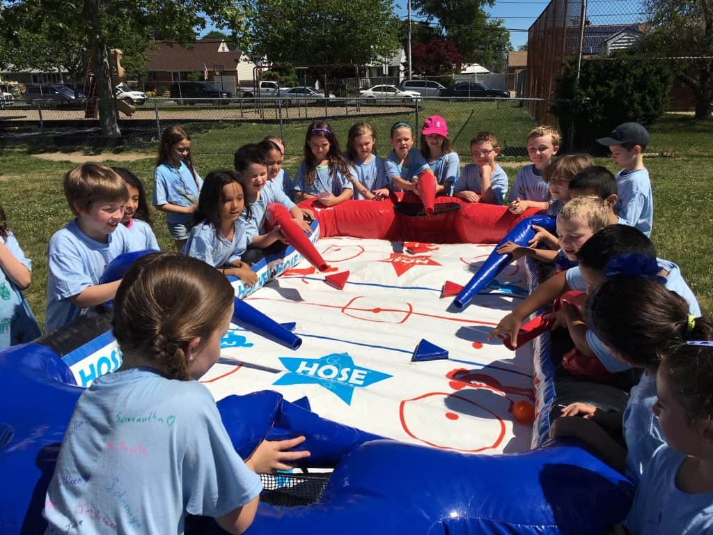 A group of children in matching blue shirts play a large, inflatable tabletop hockey game—perfect for party rentals Long Island—outdoors on a sunny day. They are gathered around the game, smiling and focusing on the play.