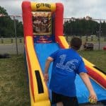 A boy in a blue shirt with the number 16 throws a ball at a colorful inflatable toss game, part of party rentals Long Island, outdoors on a grassy field with a fence and trees in the background.