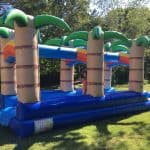 An inflatable slip-and-slide with palm tree designs stands on grass, perfect for party rentals Long Island. A child in a swimsuit is enjoying the slide outdoors on a sunny day, surrounded by trees and greenery.