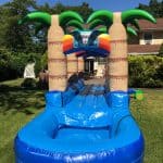 A blue inflatable slip and slide with palm tree decorations sits on green grass in a NY backyard, with a house and trees in the background under a clear blue sky—perfect for party rentals Long Island fun.