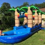 A child in a swimsuit sits at the end of a blue inflatable water slide with palm tree decorations, part of fun party rentals Long Island, in a grassy backyard on a sunny day.