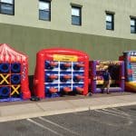 Three colorful inflatable carnival games, perfect for party rentals Long Island, NY, are set up on a sidewalk against a green building. A person stands beside the middle game among tic-tac-toe, ball toss, and a knock-off challenge.