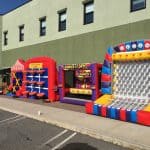 Three colorful inflatable carnival games—including tic-tac-toe, a Nerf ball toss, and a plinko-style game—are set up outdoors on a paved area next to a green building. Perfect for NY events or party rentals Long Island celebrations!.