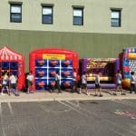 Children walk in a line past four colorful inflatable carnival games, including tic-tac-toe, basketball, knock-it-off, and ball toss—perfect for party rentals Long Island, NY—set up outside on a sunny day.