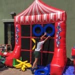 A child places a blue ring on a large inflatable tic-tac-toe game outdoors, with bounce house Long Island vibes and colorful pieces scattered nearby. The inflatable structure features red, white, and black—perfect for party rentals Long Island fun!.