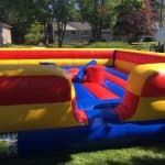 A colorful inflatable jousting arena with red, yellow, and blue sections sits on grass in a suburban yard—perfect for party rentals Long Island. Two padded jousting sticks rest in the center, surrounded by trees and houses.