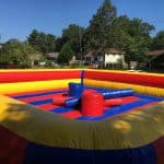 An inflatable jousting arena in red, yellow, and blue is set up outdoors with foam pedestals and padded poles—perfect for party rentals Long Island. Houses and trees make a lively backdrop for endless fun.