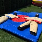 Two large padded sumo wrestling suits and helmets lie flat on a blue and red mat outdoors on grass, with greenery and bounce house Long Island equipment in the background—perfect for NY party rentals Long Island events.