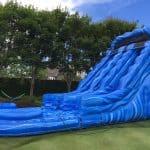 A large blue inflatable water slide with wave patterns stands on grass, perfect for party rentals Long Island, NY. Water flows down two lanes, with trees and a hedge in the background under a clear blue sky.