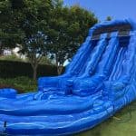 A large inflatable blue water slide with two lanes and a wavy pattern stands on a grassy lawn, surrounded by trees and shrubs under a clear blue sky—perfect for party rentals Long Island celebrations.