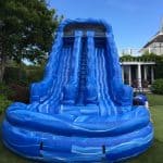 A large blue inflatable water slide with two sliding lanes and a central ladder stands on a grassy lawn, perfect for party rentals Long Island, surrounded by trees and a house in the background.
