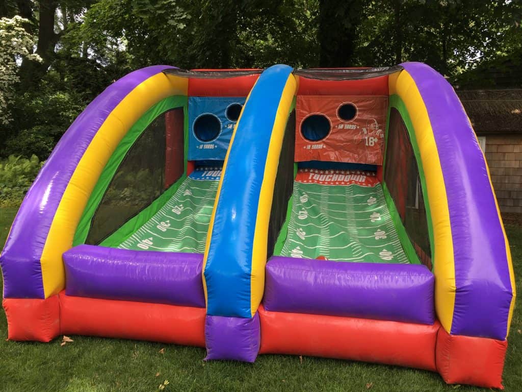 A colorful inflatable football toss game with two lanes, each featuring a target hole and yard line markings, set outdoors on green grass—perfect for NY party rentals Long Island events with trees in the background.