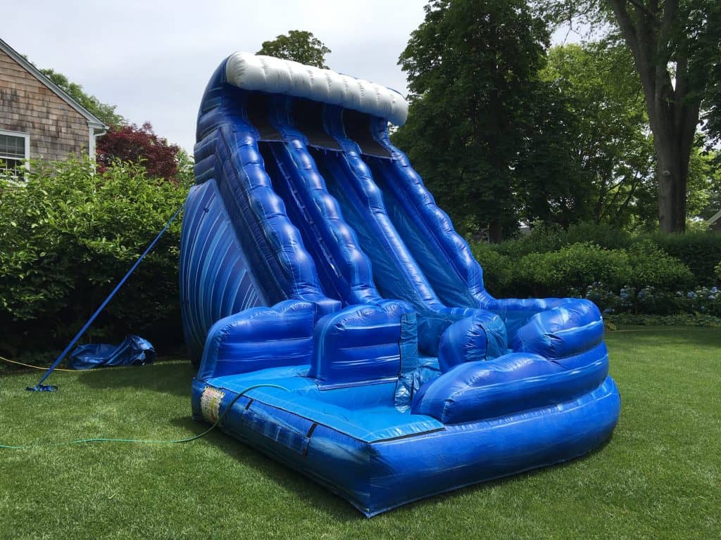 A large blue inflatable water slide shaped like a wave is set up on green grass in a backyard surrounded by bushes and trees—perfect for NY party rentals Long Island fun.