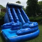 A large blue inflatable water slide with two sliding lanes and wave designs, available from party rentals Long Island, set up on green grass in a backyard with trees and shrubs in the background.