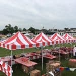 Red and white striped tents cover tables filled with snacks and treats at an outdoor event on a grassy lawn. In the background, party rentals Long Island like chairs and a bounce house add to the festive setup as people prepare for the celebration.