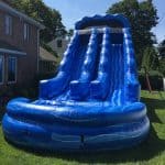 A large blue inflatable water slide with two lanes, available from party rentals Long Island, is set up on a grassy lawn next to a brick house on a sunny day.