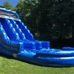 A large, blue inflatable water slide with wavy patterns sits on a grassy lawn, surrounded by trees and a sunny sky—perfect for party rentals Long Island, NY. The slide features two lanes and a shallow pool at the bottom.