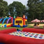 A grassy outdoor area with colorful inflatable games, including a bounce house Long Island families love, an obstacle course, and a large Twister mat. Two red-and-white striped tents are set up nearby, surrounded by trees.