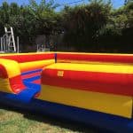 A large, inflatable, rectangular bounce house with red, yellow, and blue stripes is set up on a grassy lawn with trees and a fence in the background—perfect for party rentals Long Island, NY.