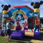 A colorful Mickey Mouse-themed bounce house Long Island with images of Mickey, Minnie, Goofy, Donald, and Daisy stands on a lawn. Two adults and a child are near the entrance at this sunny NY party rentals Long Island event.