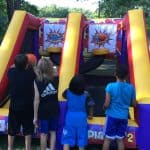 Four children play outdoors on an inflatable basketball arcade game, aiming for two hoops. With trees and grass in the background, it’s a perfect scene for party rentals Long Island, NY—fun like a bounce house Long Island kids love!.