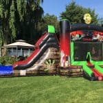 Large inflatable bounce house and slide with a zombie theme, featuring green and red colors, monster graphics, and a skull at the top—perfect for NY parties or anyone seeking party rentals Long Island—set up on a grassy lawn with trees and a gazebo nearby.